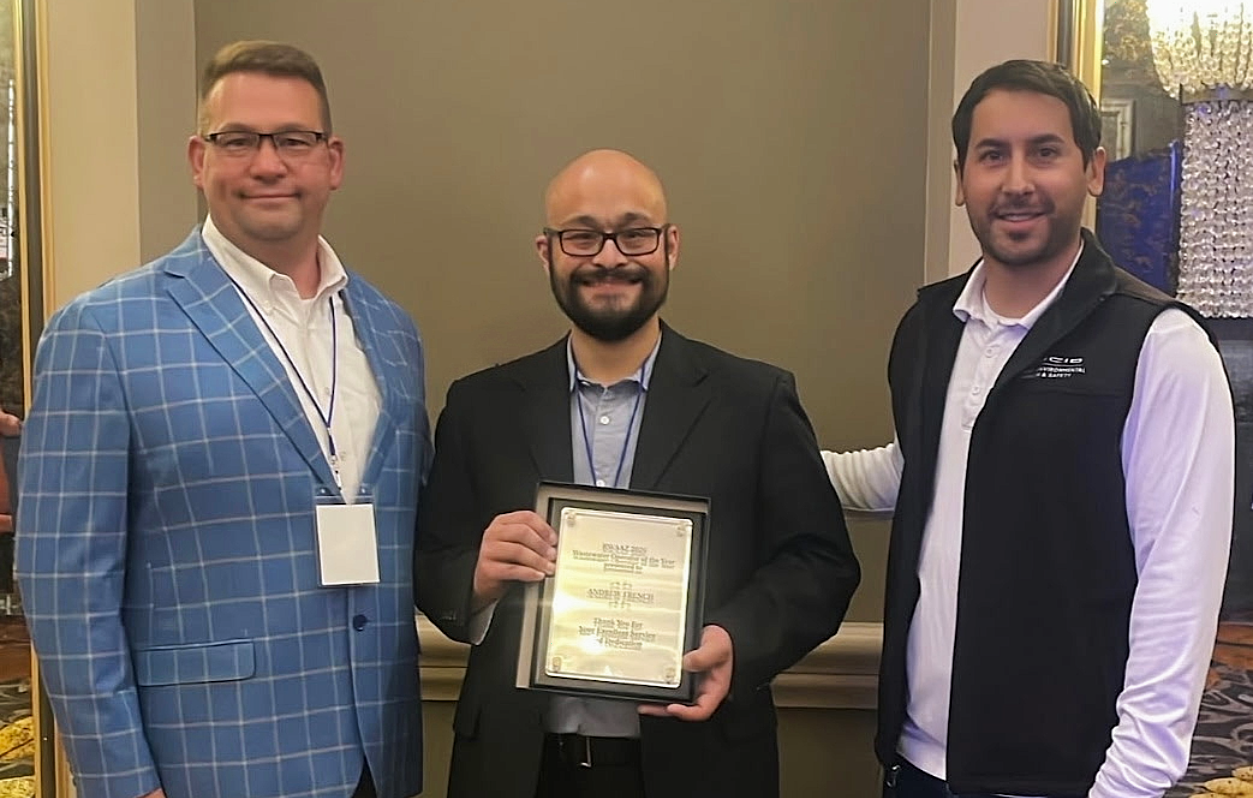 Justin Niles (left), Andrew French (center) and a representative from a customer (right) standing indoors at the Annual Rural Water Association conference. Andrew French holds a framed plaque recognizing Wastewater Operator of the year. All are wearing business or business-casual attire, and the background includes neutral walls and decorative lighting.