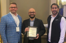 Justin Niles (left), Andrew French (center) and a representative from a customer (right) standing indoors at the Annual Rural Water Association conference. Andrew French holds a framed plaque recognizing Wastewater Operator of the year. All are wearing business or business-casual attire, and the background includes neutral walls and decorative lighting.