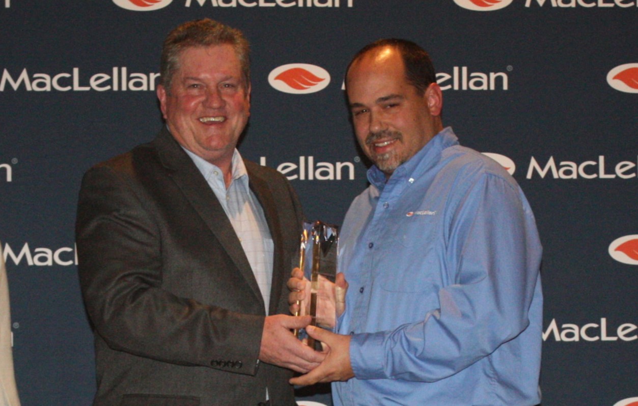 John Evans (left) presenting the engraved cyrstal leadership award to Robby Clendening (right) in front the the photo backdrop with the MacLellan logo repeated across it. 